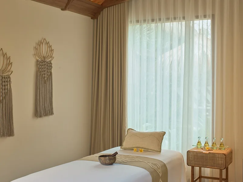 A massage table with towels and small bottles on a side table beside a window with sheer curtains